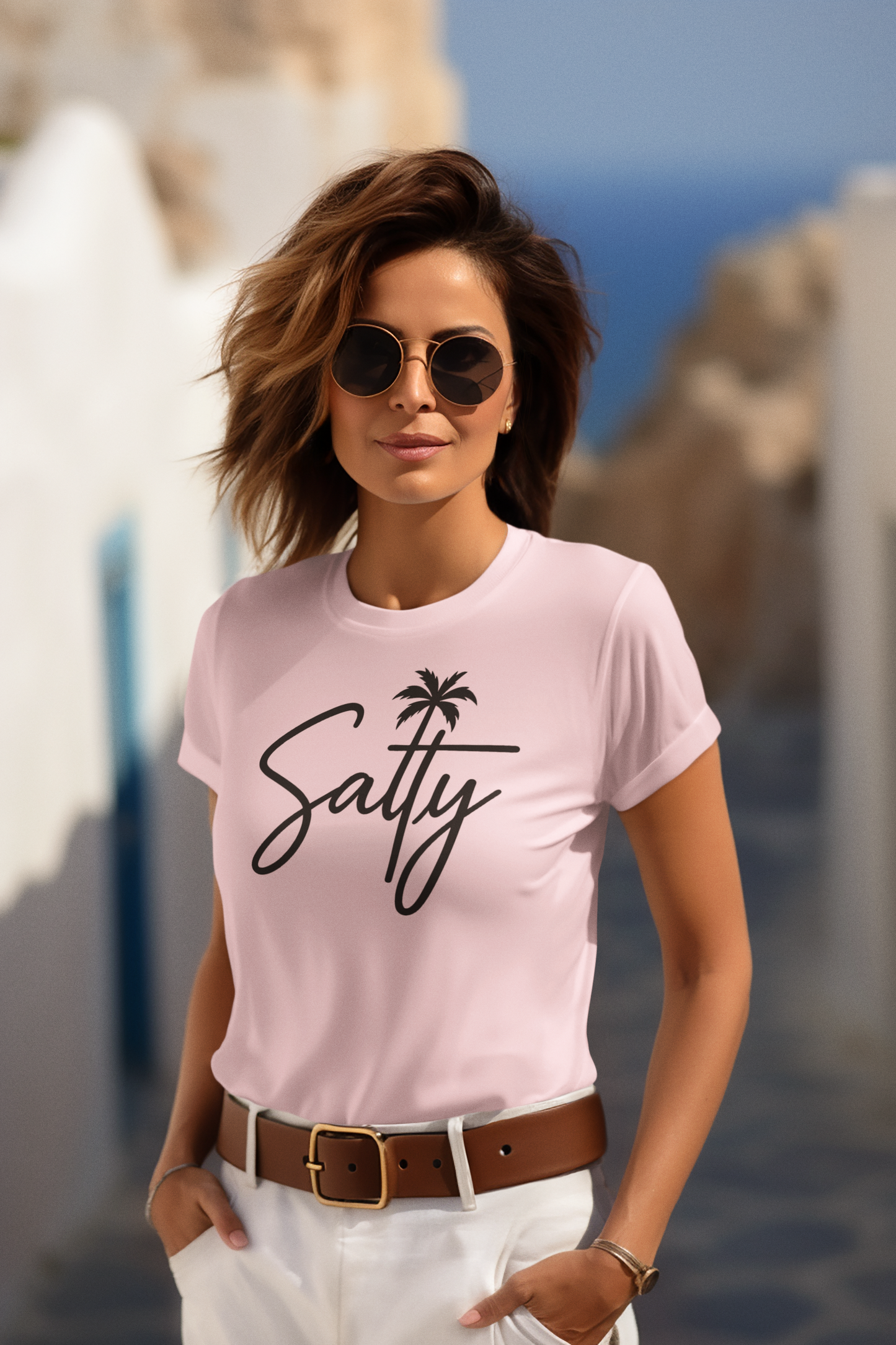 Woman wearing a pink t-shirt with 'Salty' text and palm tree design, sunglasses, and white pants in a scenic location.