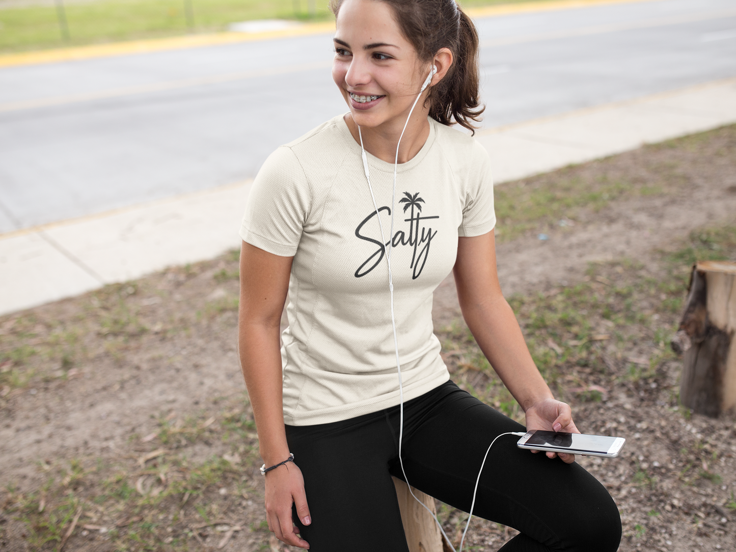 Woman wearing a 'Salty' t-shirt, sitting outdoors with earphones and a phone.