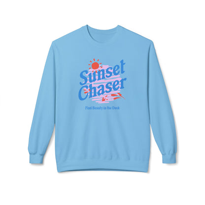 A light blue crewneck sweatshirt featuring a retro-style graphic on the chest with the text 'Sunset Chaser' in blue and red, a small red sun, and the phrase 'Find Beauty in the Dark' underneath, displayed on a clean white background.