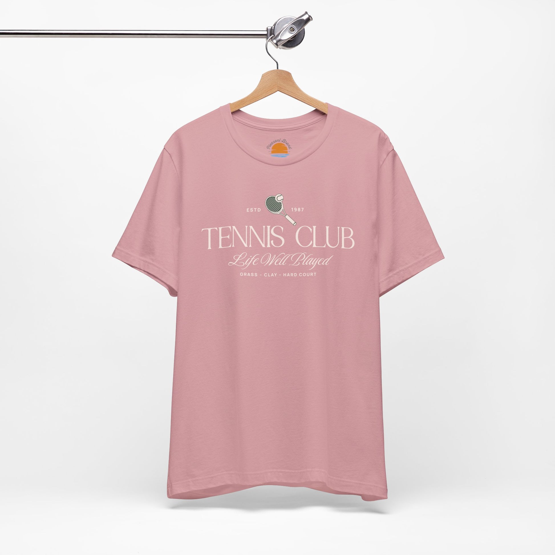 Pink t-shirt with 'Tennis Club' text and graphics on a hanger against a white background