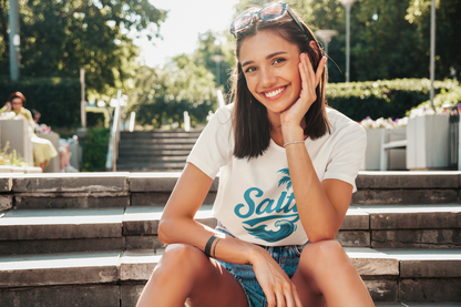 Woman sitting on steps outdoors wearing a t-shirt with 'Salty' blue wave and palm tree design 
