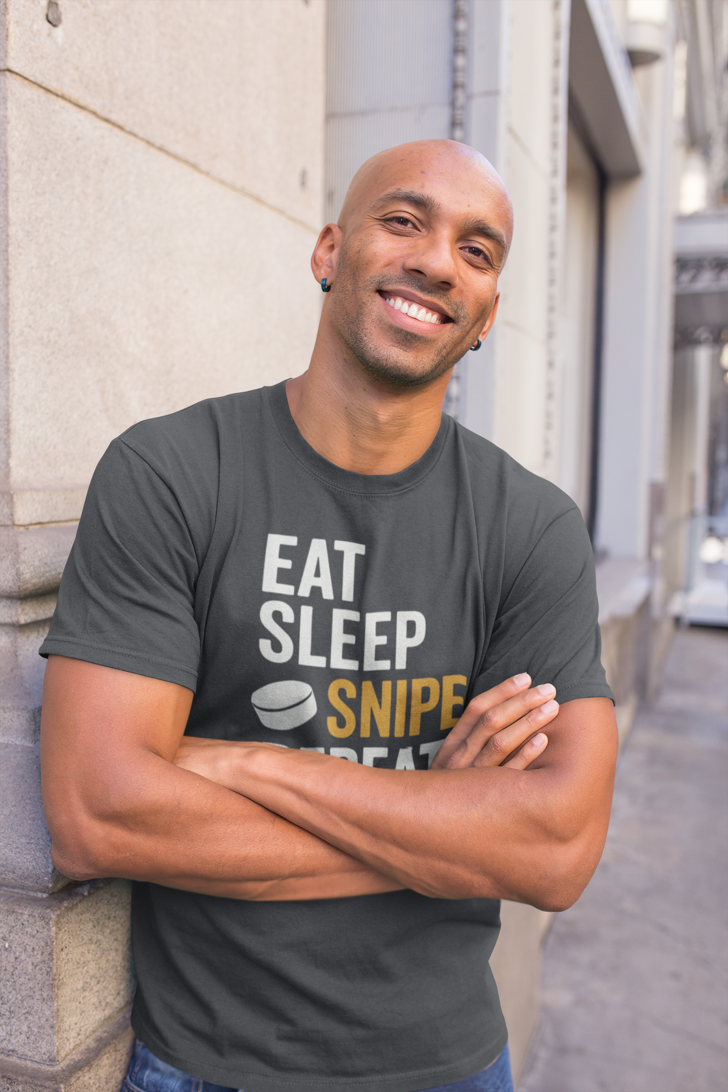Confident man wearing Eat Sleep Snipe Repeat T-Shirt with arms crossed showing scorer swagger, standing against a building.