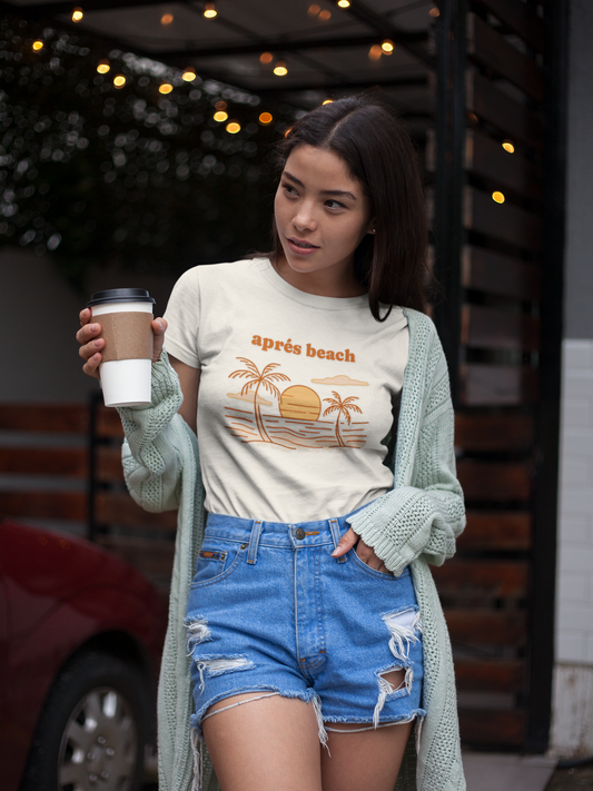 Woman wearing a natural color 'après beach' sunset and palm trees graphic t-shirt holding a coffee cup outdoors.