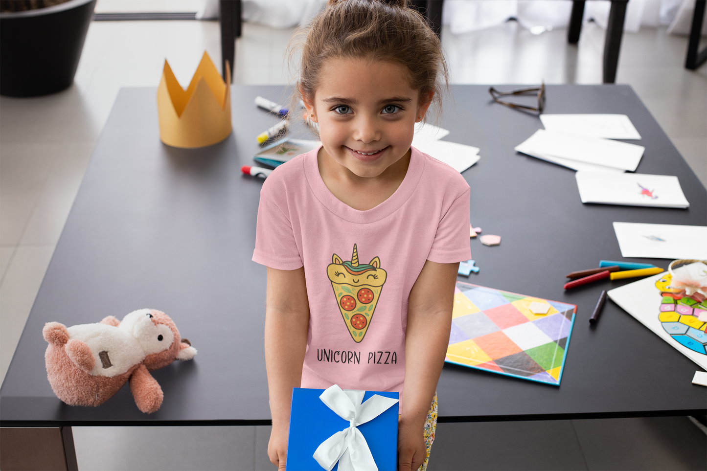 Smiling child in pink Unicorn Pizza T-Shirt surrounded by colorful art supplies and projects