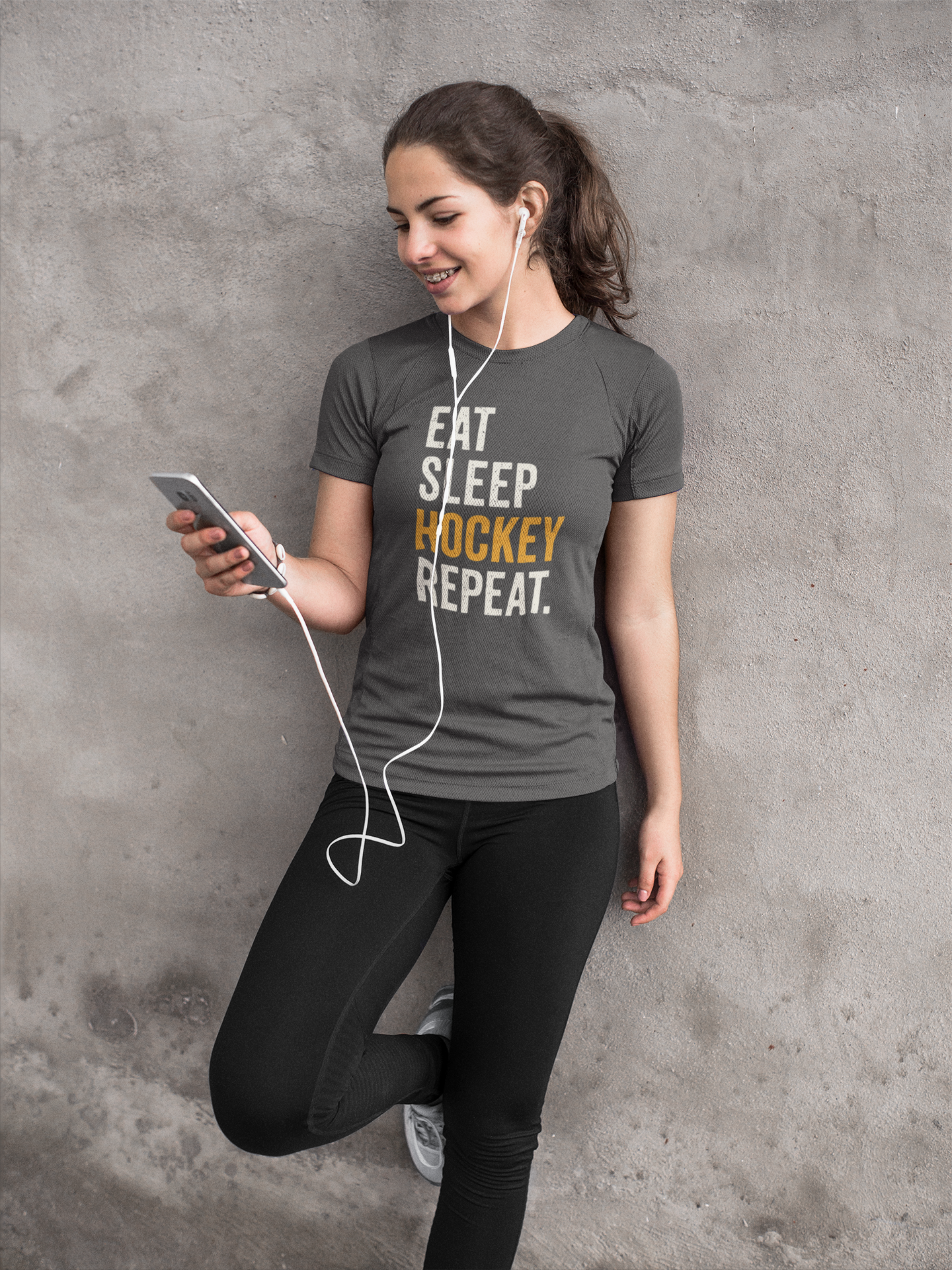 Smiling teen in Eat Sleep Hockey Repeat Youth T-Shirt in relaxed lifestyle pose with phone and earbuds
