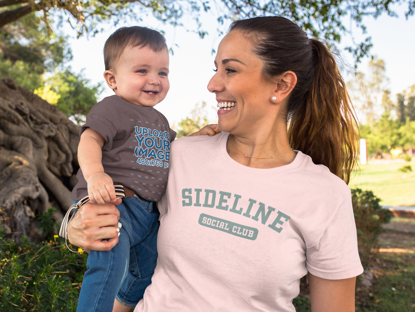 Woman holding a child outdoors with 'SIDELINE SOCIAL CLUB' t-shirt