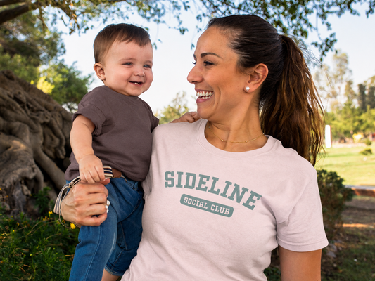 Woman holding a child outdoors with 'SIDELINE SOCIAL CLUB' t-shirt