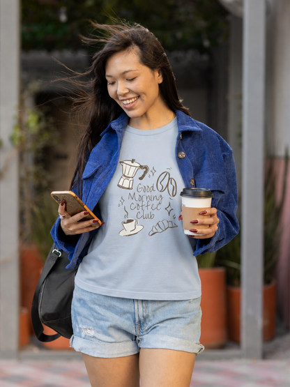 Woman walking outdoors, holding a coffee cup and phone, wearing a blue t-shirt with a coffee-themed graphic design.