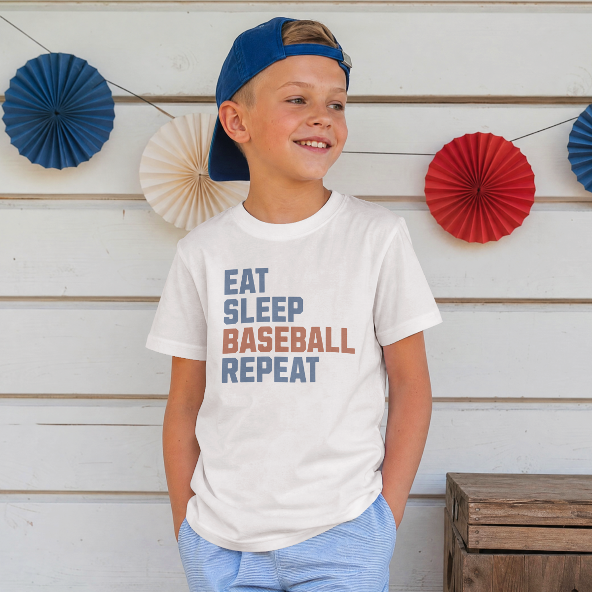 Child wearing a white t-shirt with 'Eat Sleep Baseball Repeat' text, standing against a wooden wall with decorative fans.