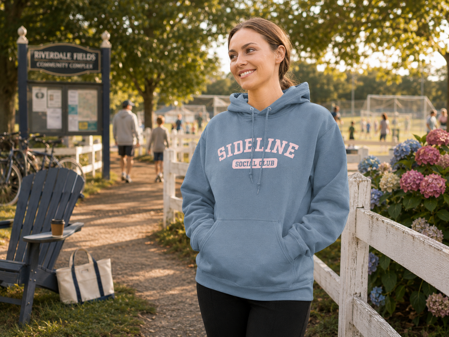 A woman wearing a dusty blue hoodie with 'SIDELINE SOCIAL CLUB' printed on the front, standing outdoors in a park-like setting with trees, a fence, and a sign in the background.