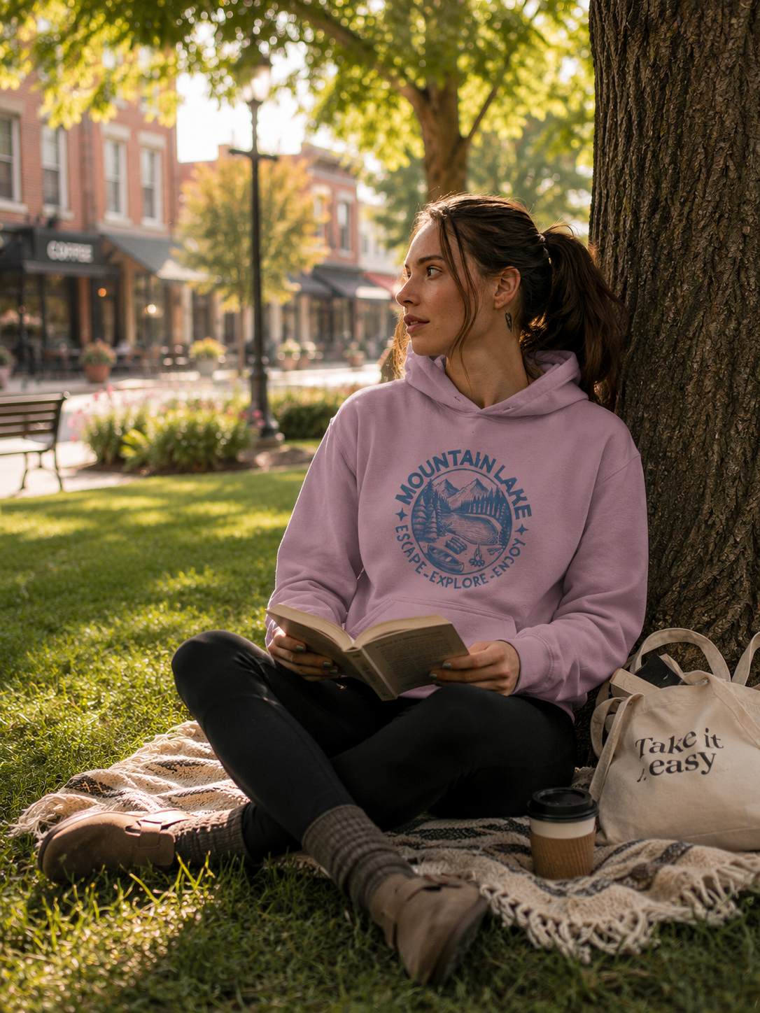 A woman sits on a blanket on the grass in a park, reading a book. She is wearing a light purple hoodie with a blue 'Mountain Lake Escape-Explore Enjoy' graphic printed on the chest, black leggings, and brown boots.