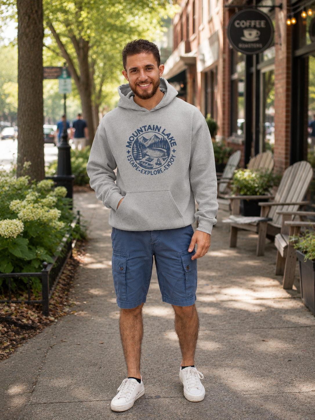 A man standing on a suburban downtown sidewalk wearing a light gray pullover hoodie with a dark blue'Mountain Lake Escape-Explore Enjoy' graphic printed on the chest, paired with blue cargo shorts and white sneakers.