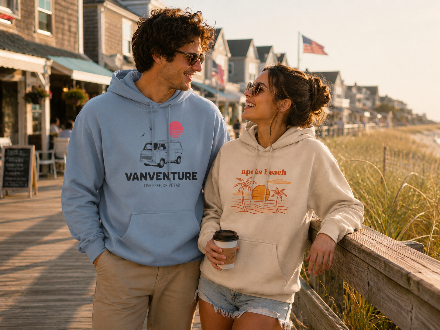A man and a woman standing on a wooden boardwalk by the beach, wearing hoodies. The man wears a light blue hoodie with a camper graphic and 'VANVENTURE' text, while the woman wears a cream hoodie with an orange beach graphic and 'apres beach' text.