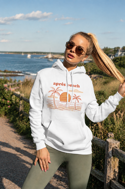 A woman wearing a white pullover hoodie with a retro orange beach graphic and the text 'apres beach' stands outdoors near a wooden fence and water.