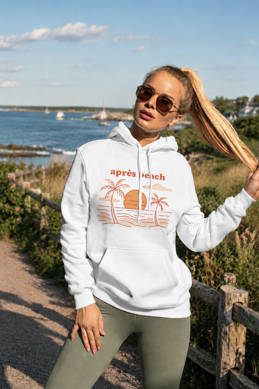A woman wearing a white pullover hoodie with a retro orange beach graphic and the text 'apres beach' stands outdoors near a wooden fence and water.