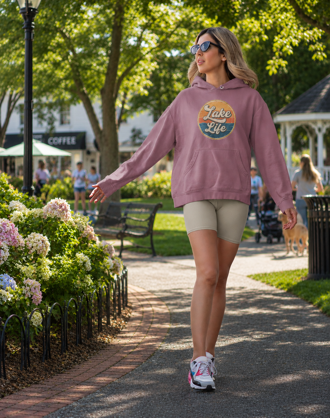 A woman walking on a park path wearing a dusty rose hoodie with a 'Lake Life' graphic on the chest, paired with beige biker shorts and white sneakers.