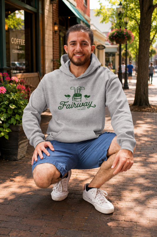 A man crouching on a brick sidewalk in front of a storefront, wearing a light gray hoodie with a green 'Fairway' graphic and blue shorts.