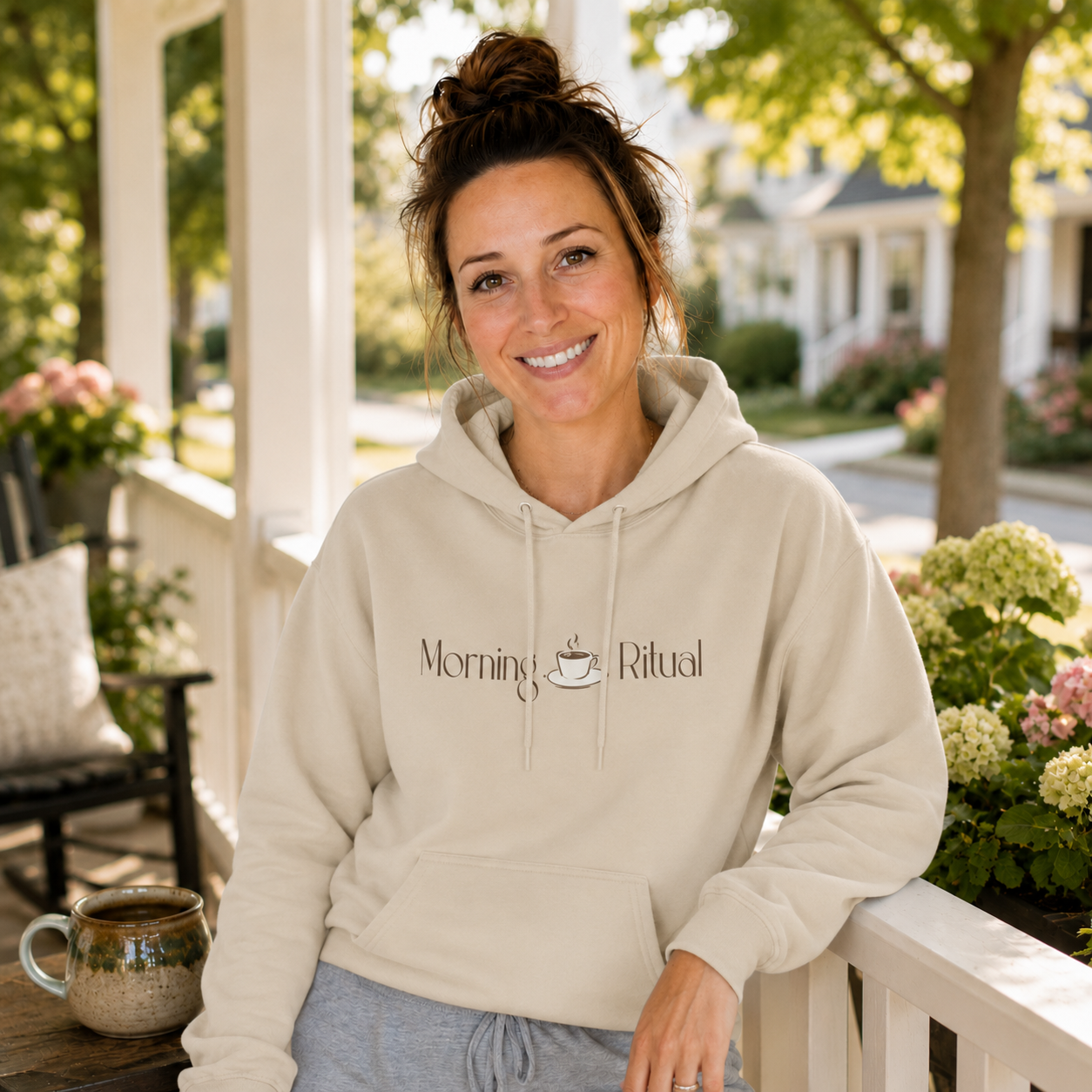 A smiling woman with her hair in a bun sits on a white porch railing wearing a beige hoodie with the words 'Morning Ritual' and a small coffee cup graphic. A mug sits on a table beside her, with a sunny, blurred suburban background.
