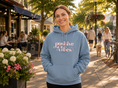 A woman wearing a light blue hoodie with 'positive vibes' printed in pink script on the front, standing outdoors on a sunny sidewalk in front of a cafe.