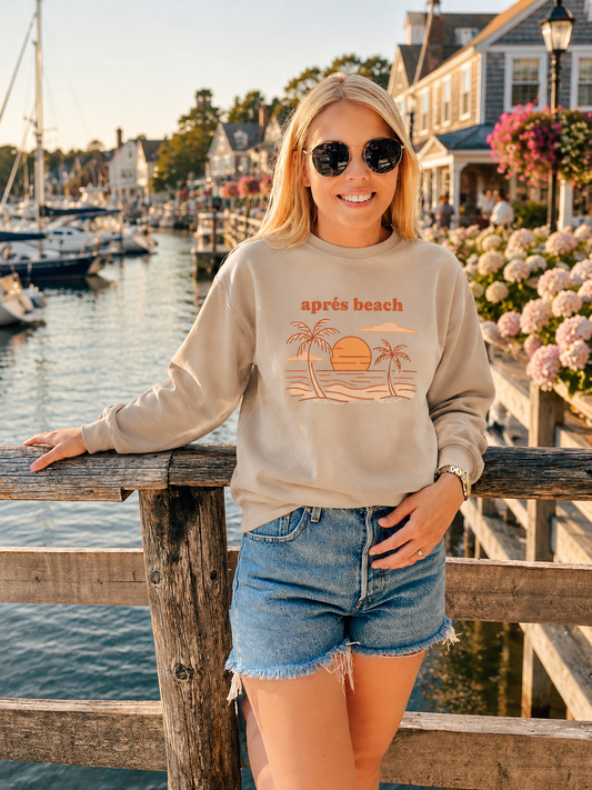 A woman wearing a beige crewneck sweatshirt with a retro beach graphic and the text 'apres beach' stands on a wooden dock by a harbor with boats and waterfront buildings in the background.
