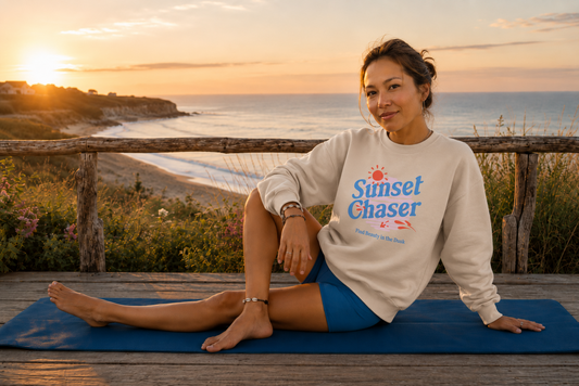 A woman sits on a blue yoga mat on a wooden deck at sunset, wearing a cream sweatshirt with a blue and red 'Sunset Chaser' graphic and blue athletic shorts, with a beach and ocean in the background.