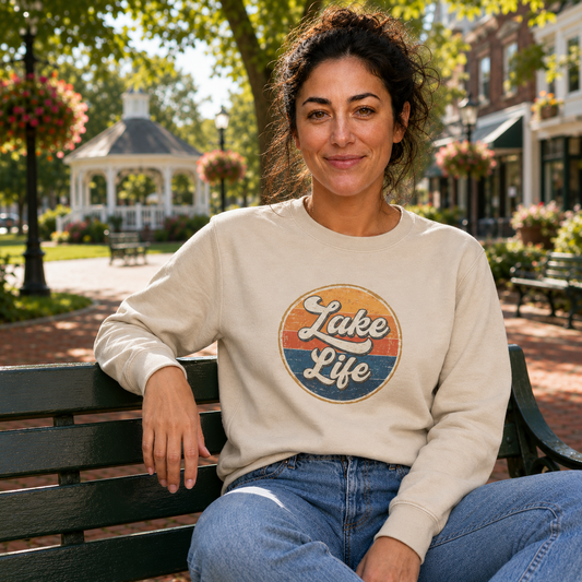 A woman sitting on a park bench wearing a beige crewneck sweatshirt with a retro circular 'Lake Life' graphic, set against a sunny, blurred background of a park with a gazebo.