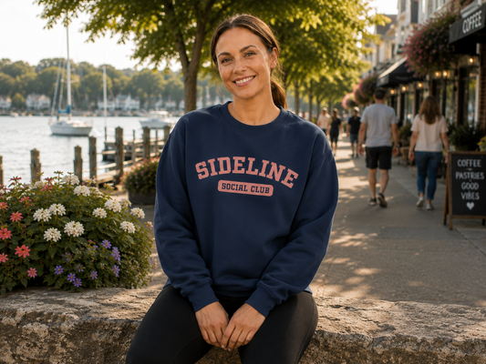 A woman sitting on a stone wall by the water wearing a navy blue crewneck sweatshirt with 'SIDELINE SOCIAL CLUB' printed in pink and white across the chest.