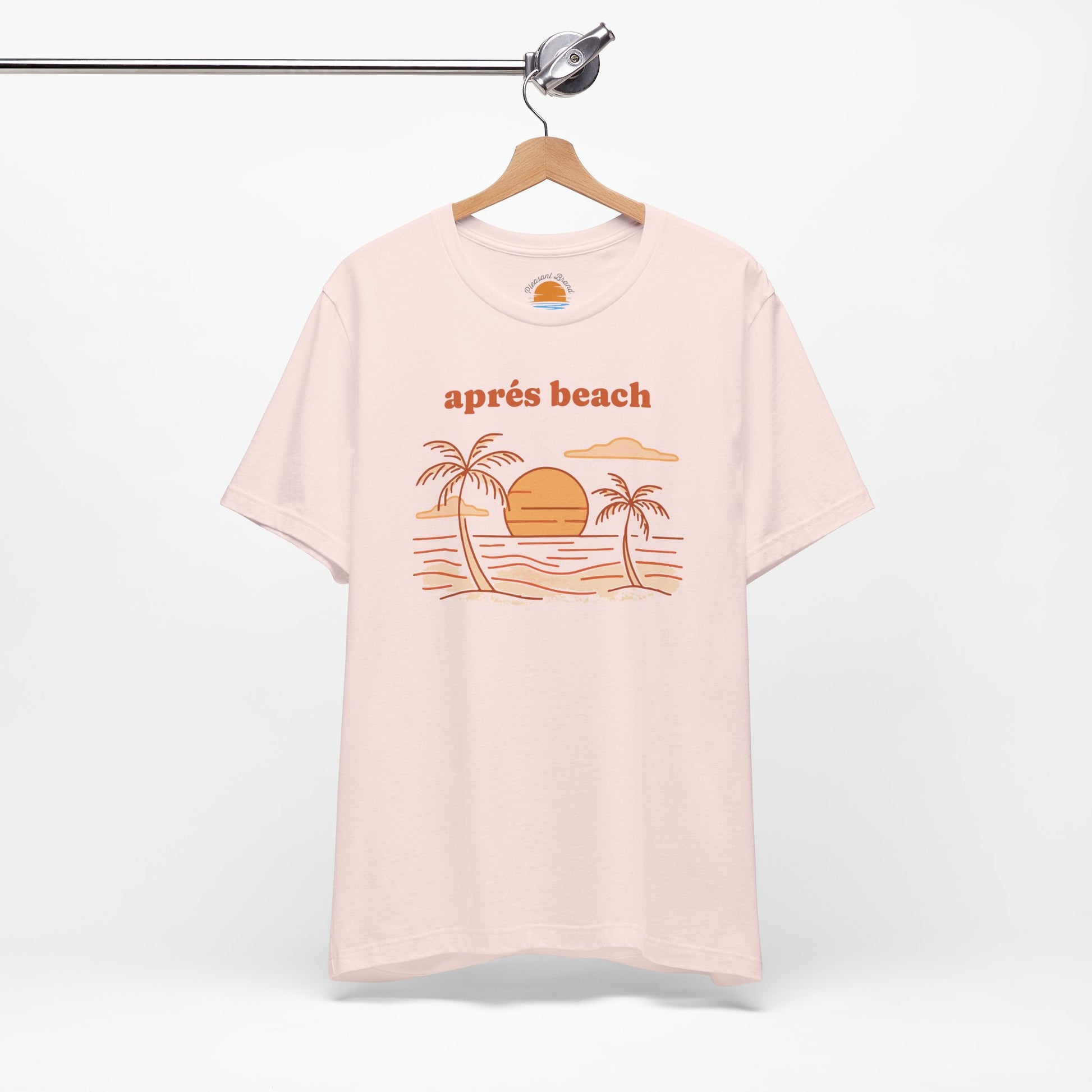 Pink color t-shirt with 'après beach' sunset and palm trees graphic design on a hanger against a white background.