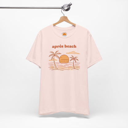 Pink color t-shirt with 'après beach' sunset and palm trees graphic design on a hanger against a white background.