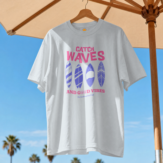 Light blue t-shirt with 'Catch Waves and Good Vibes' text and surfboard designs on a hanger under an umbrella.