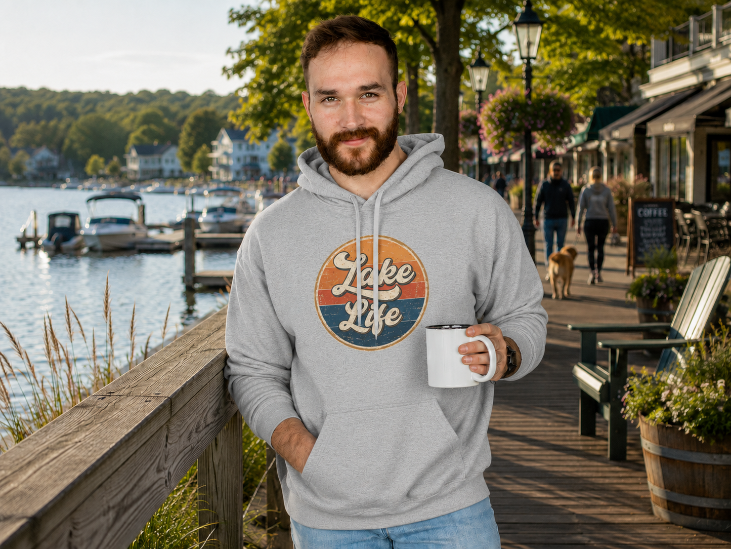 A man wearing a light gray hoodie with a retro circular 'Lake Life' graphic stands on a wooden boardwalk by the water, holding a white mug.