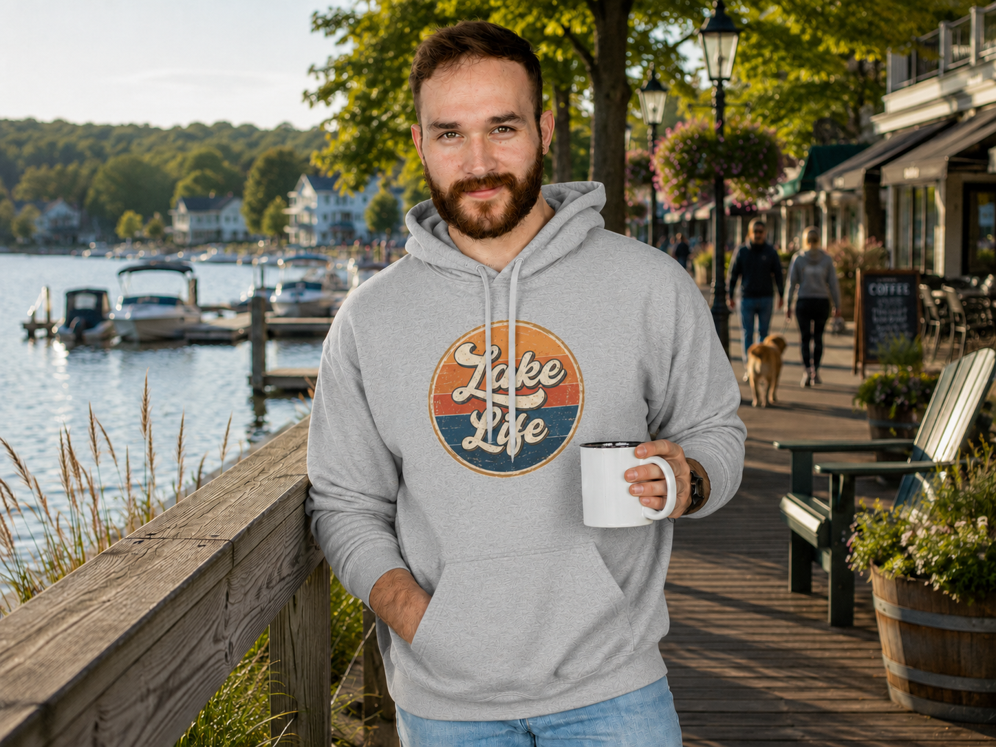 A man wearing a light gray hoodie with a retro circular 'Lake Life' graphic stands on a wooden boardwalk by the water, holding a white mug.